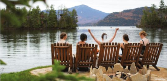 Bride and bridesmaids sitting lakeside in Adirondack chairs.
