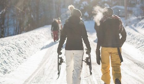 Two people walk down a snowy path, carrying snowboards. They are dressed in winter gear, including hats and gloves. Visible breath indicates cold weather. Other people are visible in the background, also walking in the snow.