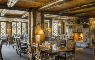 Rustic restaurant interior with wooden beams, tables, and chairs. A stone fireplace with a lit fire adds warmth. Large windows overlooking Mirror Lake allow natural light to flood in. Tables are set with plates, glasses, and cutlery. Cozy and inviting atmosphere.